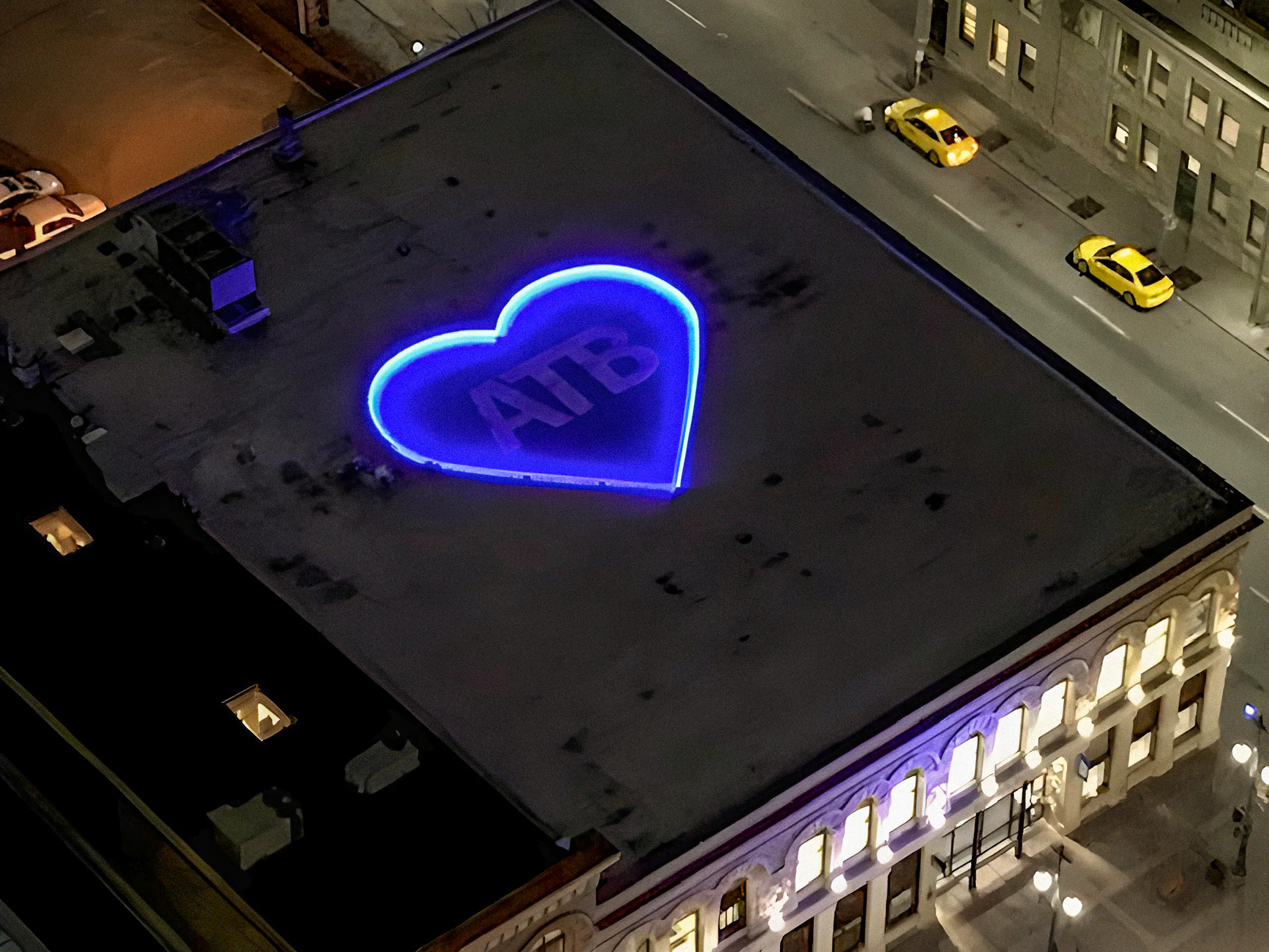 Aerial night view of rooftop with large glowing blue LED heart outline containing "ATB" letters, city streets and yellow taxis visible below