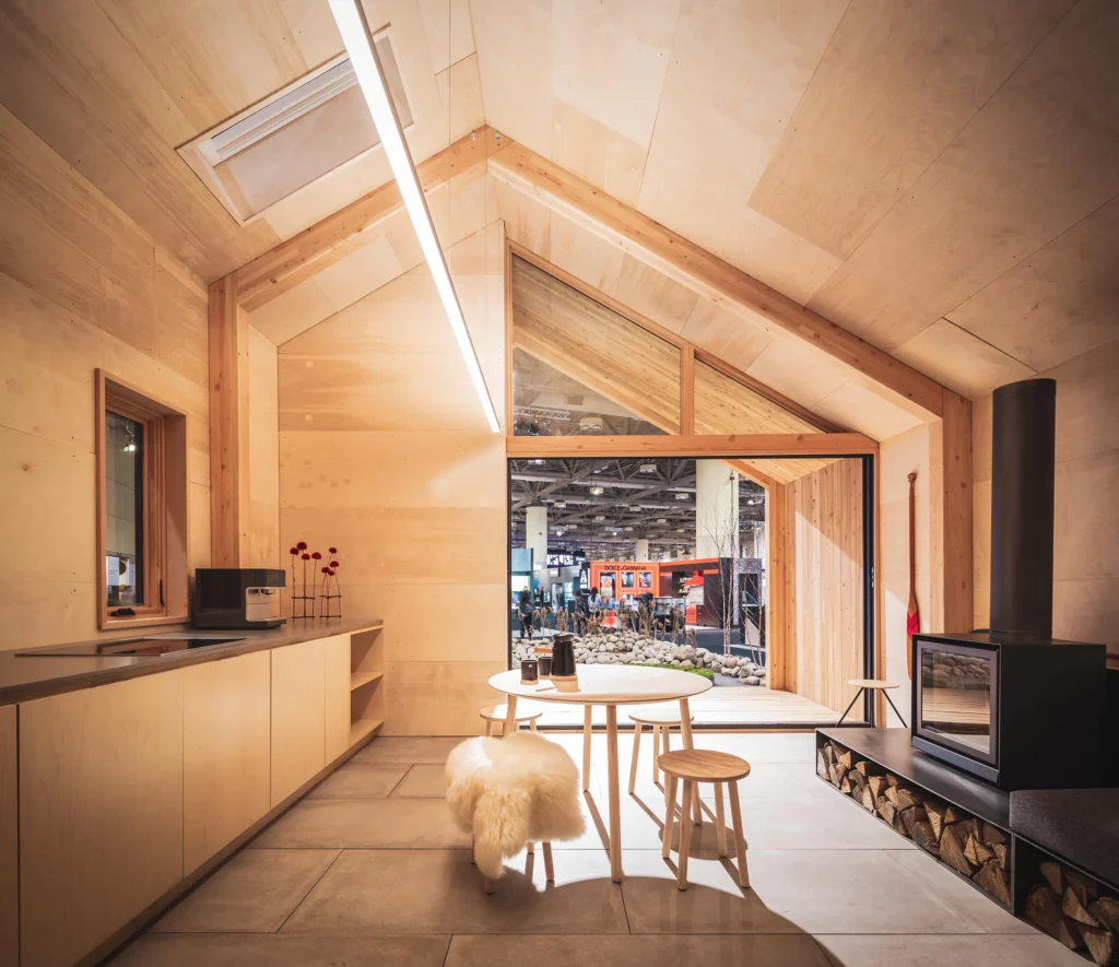 Bright modern cabin interior with vaulted cedar ceiling, linear LED lighting along the ridge, wood kitchen, round table, stove, and large glass door opening to exhibition hall