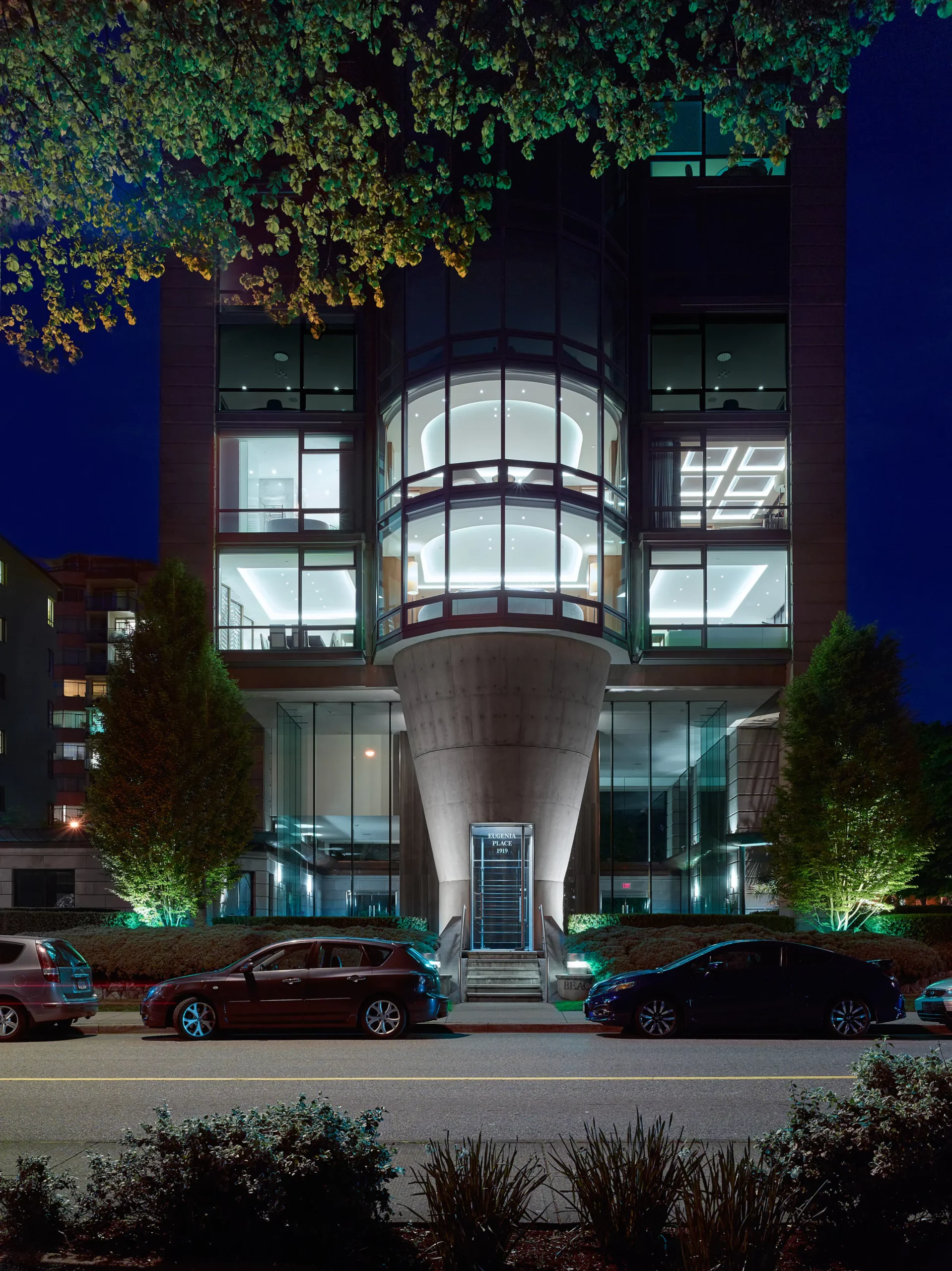 Night view of modern building with curved glass atrium illuminated by interior linear LED strips, concrete base with green uplights, entry canopy glow, and urban street foreground