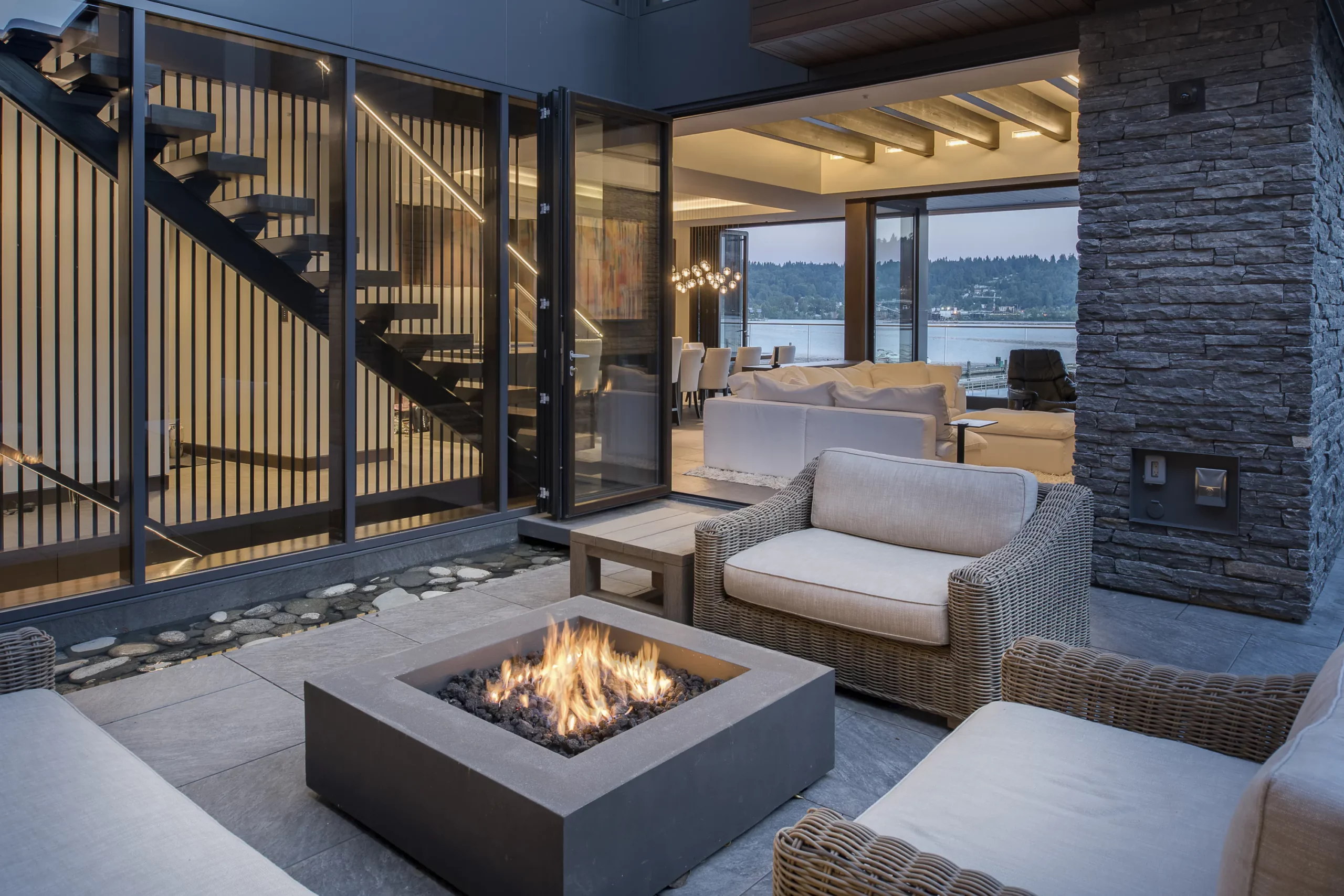 Modern patio at dusk with square fire pit flames, linear LED lighting along glass doors and stairs, rattan seating, stone cladding, and waterfront view