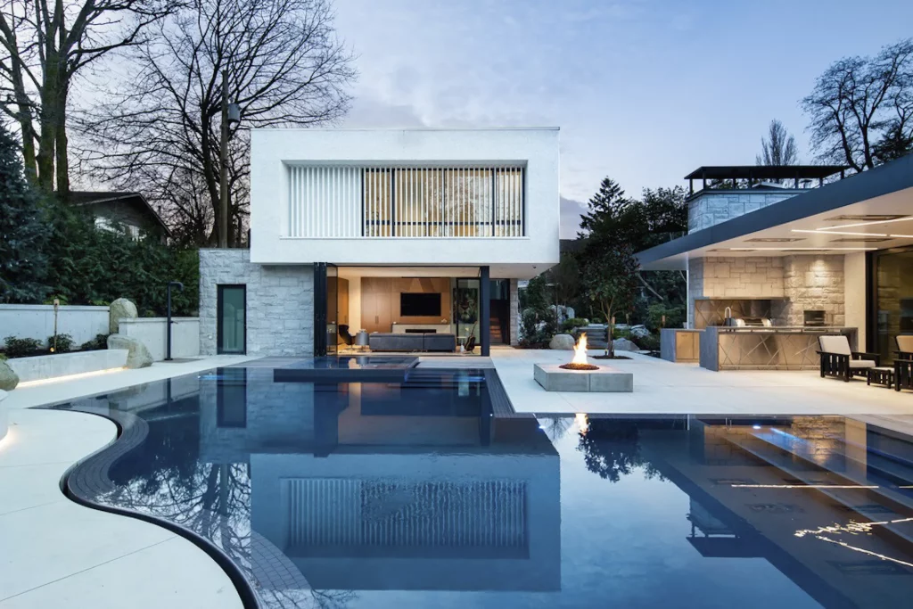 Modern luxury home exterior at dusk featuring infinity pool, central fire pit, outdoor kitchen, and linear LED architectural lighting on white facade