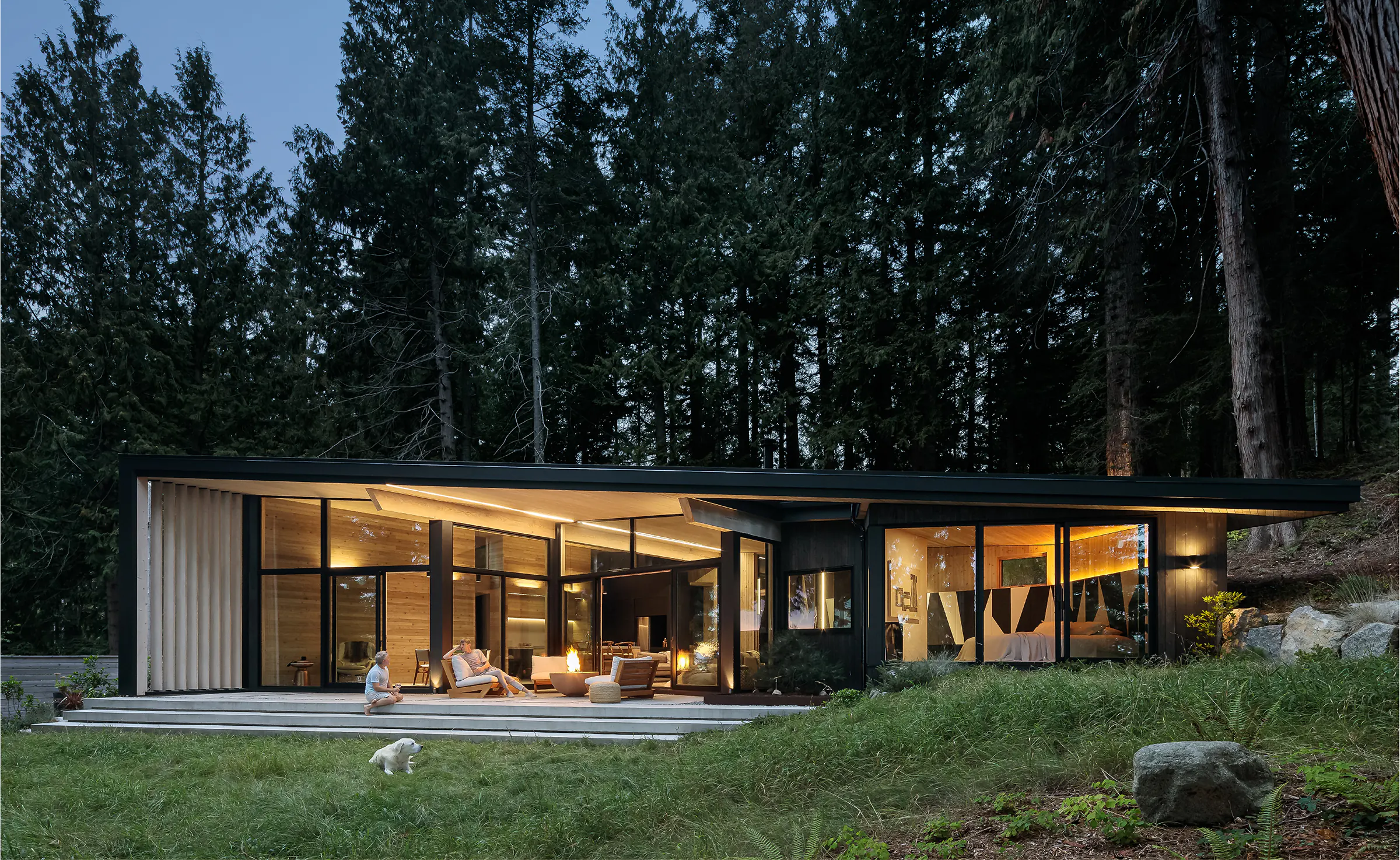 Modern cabin exterior at dusk with linear LED lighting along roof overhang, large glass walls revealing warm interior, fire pit, people relaxing, and tall forest trees