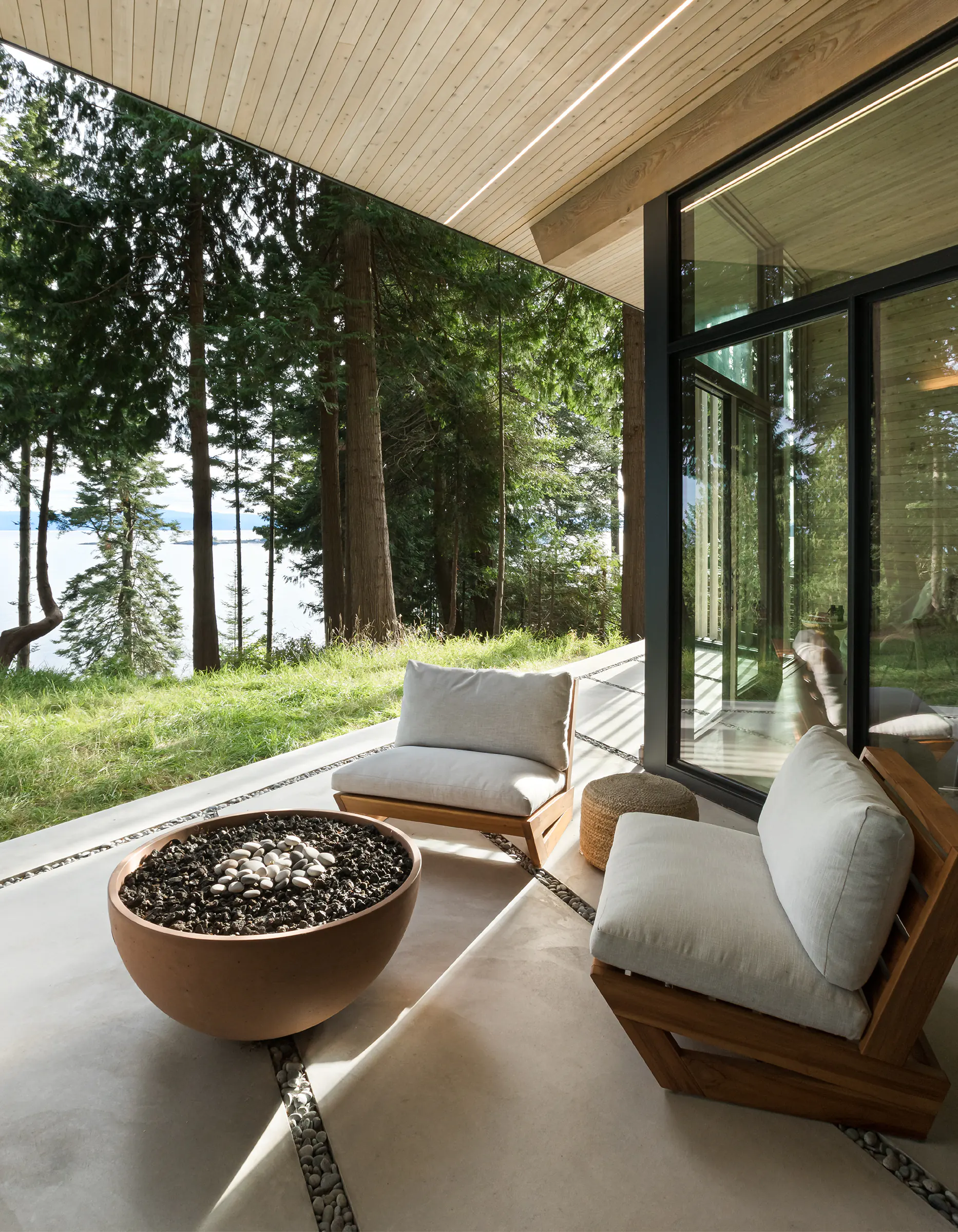 Serene modern patio at dusk with round bronze fire bowl, wooden seating with beige cushions, large glass walls to forest interior, tall evergreen trees, and waterfront view