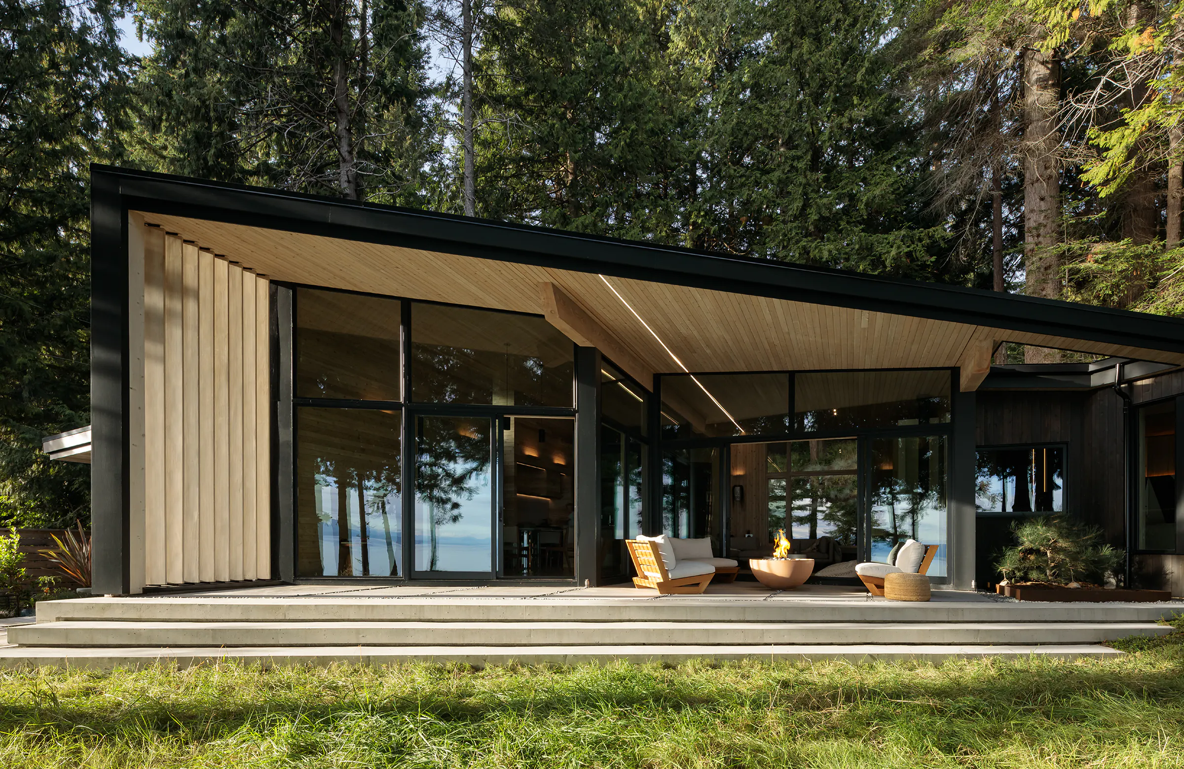 Modern cabin exterior at dusk with linear LED along roof overhang, large open glass walls showing warm interior, central fire bowl on deck, wooden seating, and tall forest trees