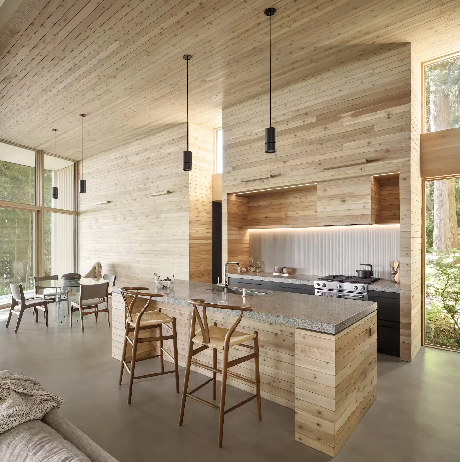 Bright modern kitchen-living interior with cedar wood walls and vaulted ceiling, linear LED strips under cabinets and along edges, large glass walls to forest, island with granite top, pendant lights, and Wishbone stools