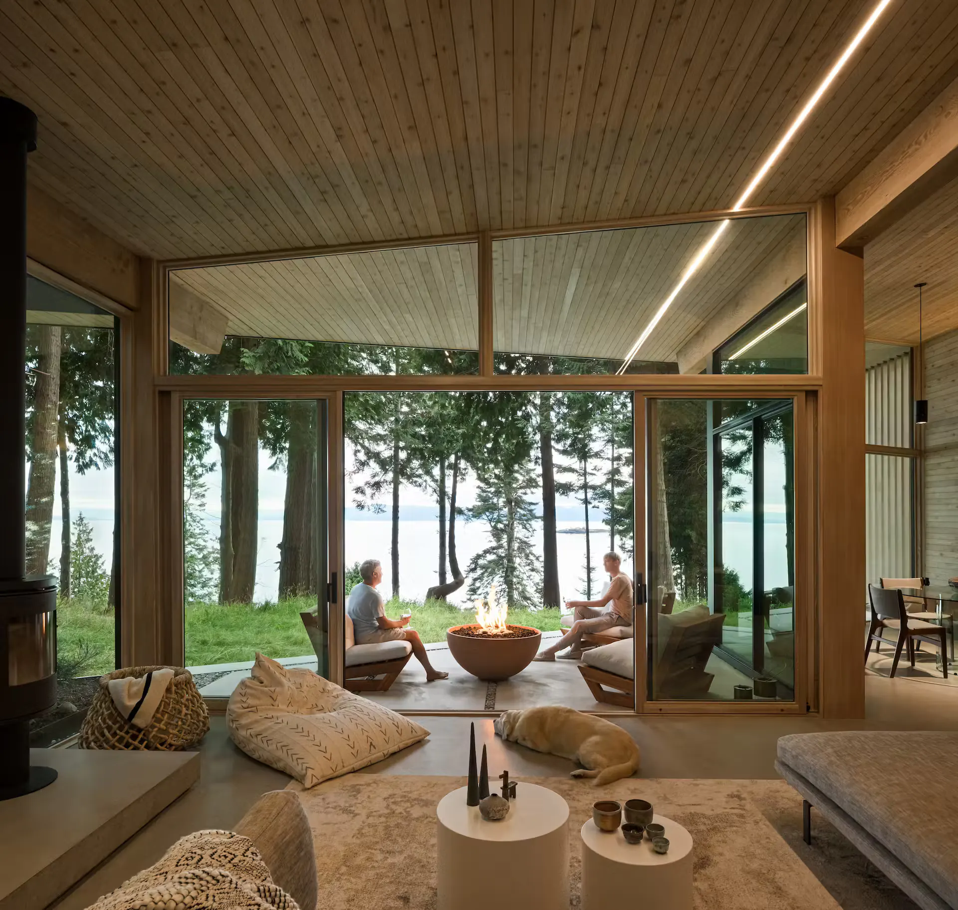 Spacious modern living area with cedar vaulted ceiling, linear LED strips glowing along roof peaks, large open glass walls to forest and water, central fire bowl, people relaxing, and dog