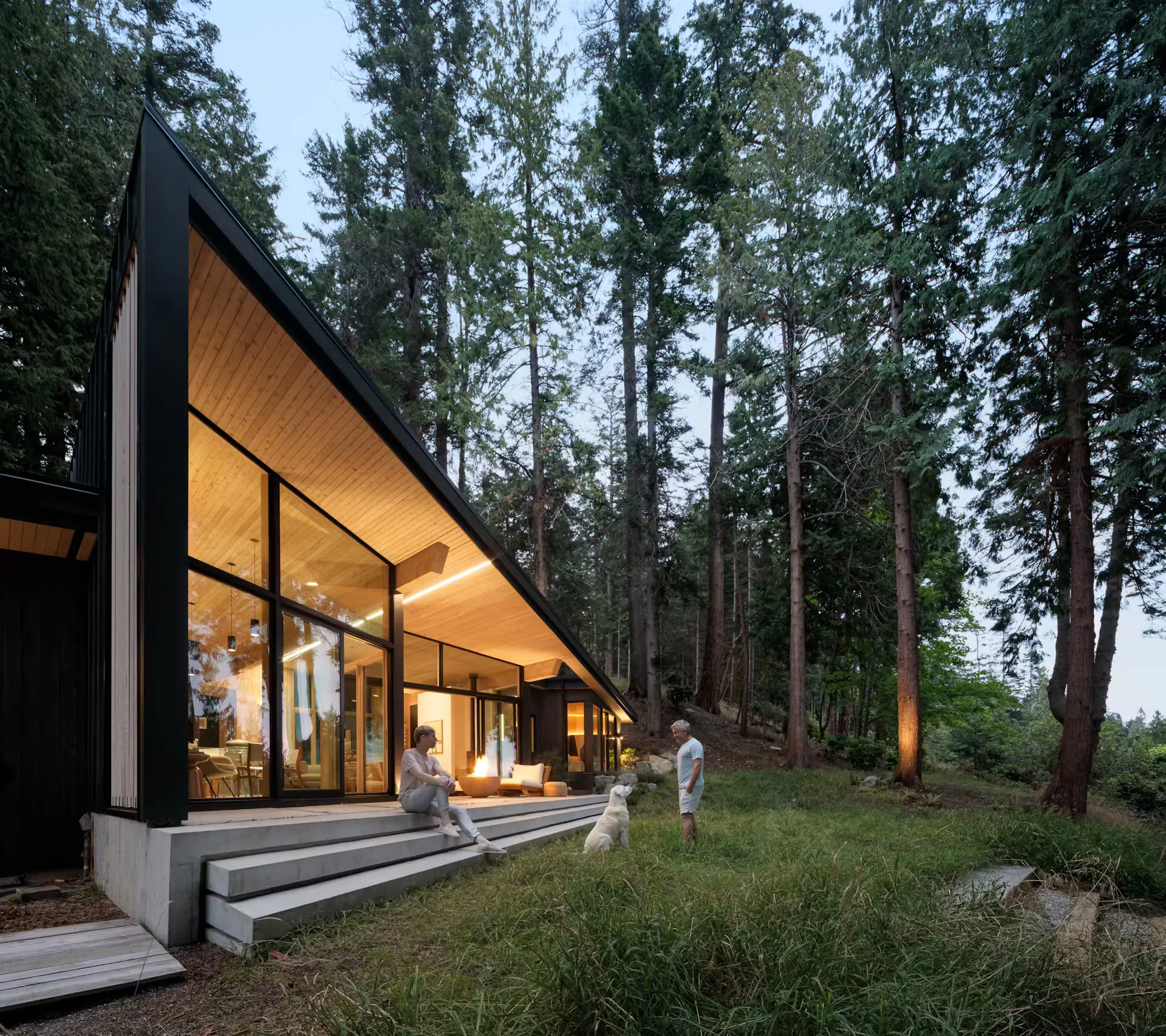 Modern cabin exterior at dusk with linear LED lighting along roof overhang, large glass walls showing warm interior, fire pit, people relaxing, dog, and tall forest trees