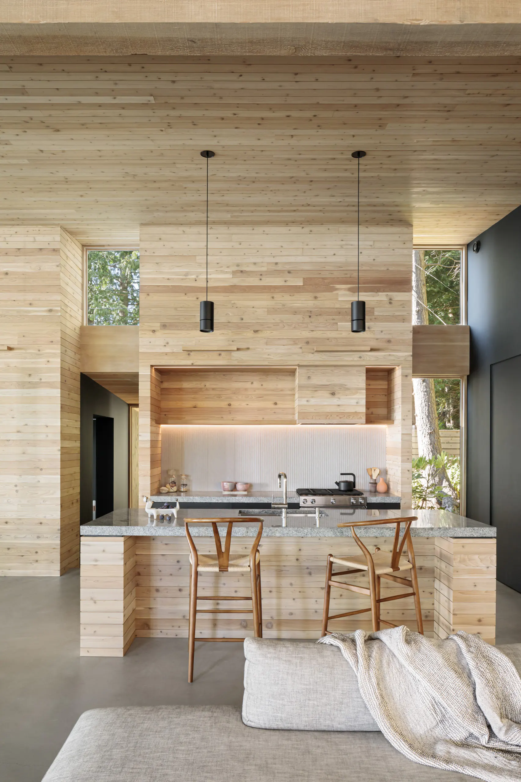 Bright modern kitchen with full cedar wood paneling, linear LED strips under cabinets and along backsplash, granite island, Wishbone stools, pendant lights, and large glass walls to forest