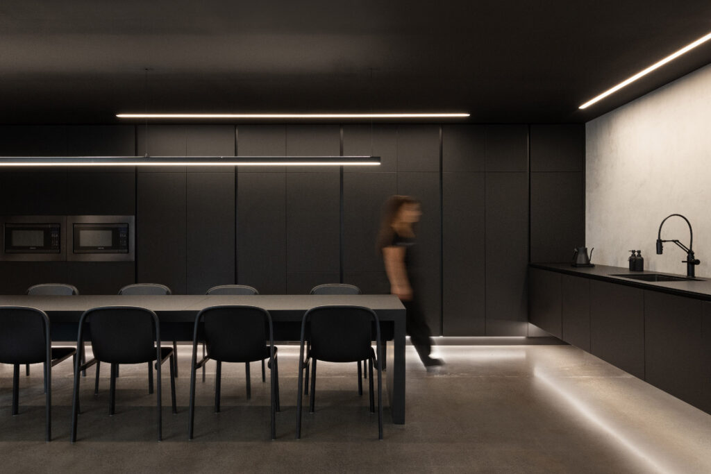 Dark modern kitchen-dining space with black cabinetry, long linear LED strips along ceiling, under-cabinet, and floor perimeter, large black table, and blurred figure walking through
