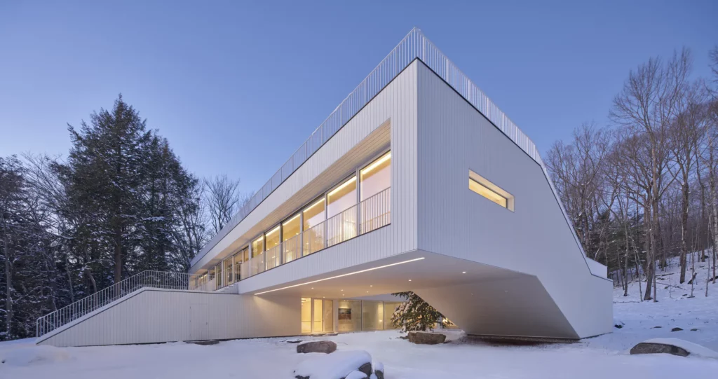 Modern angular house exterior at dusk with white corrugated cladding, long glowing window bands, cantilevered form, and snowy forest backdrop