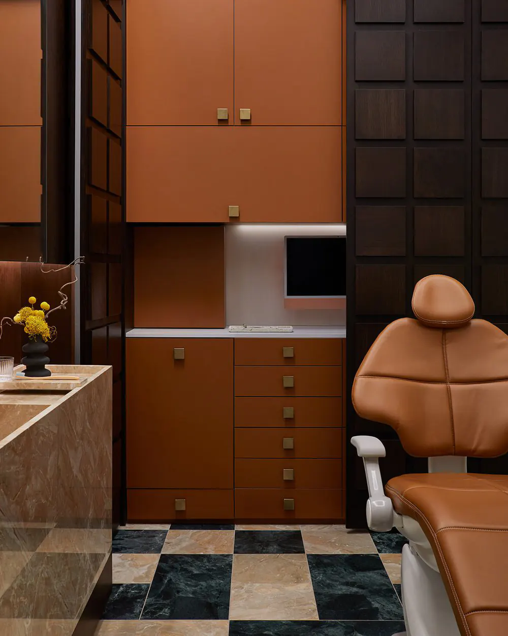 Modern dental treatment room with terracotta-orange cabinets, dark wood paneling, linear LED strips under cabinets and in niches, tan leather chair, marble countertop, and geometric marble floor