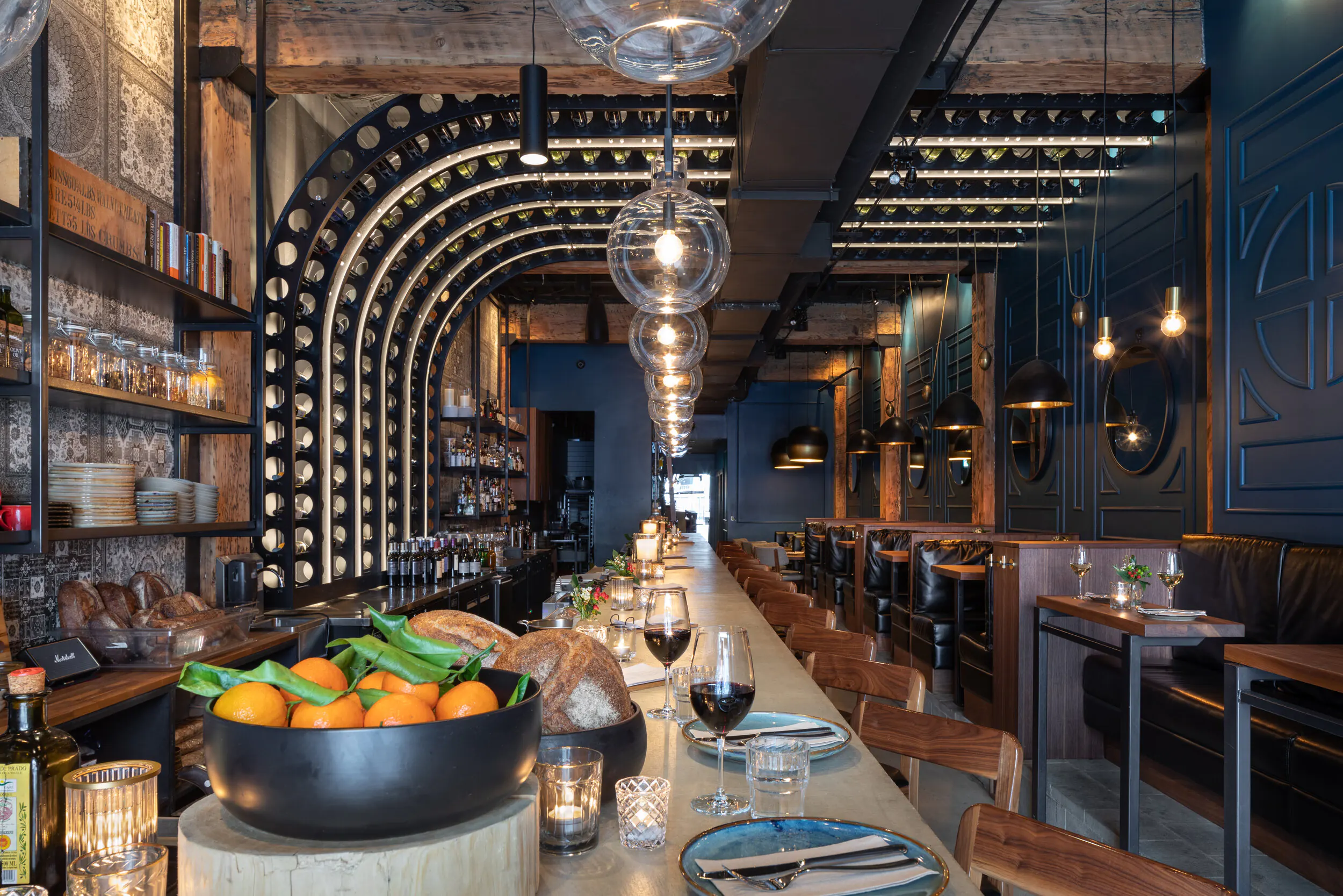 Restaurant interior featuring arched wine rack with integrated LED lighting, pendant globes, and long communal dining table