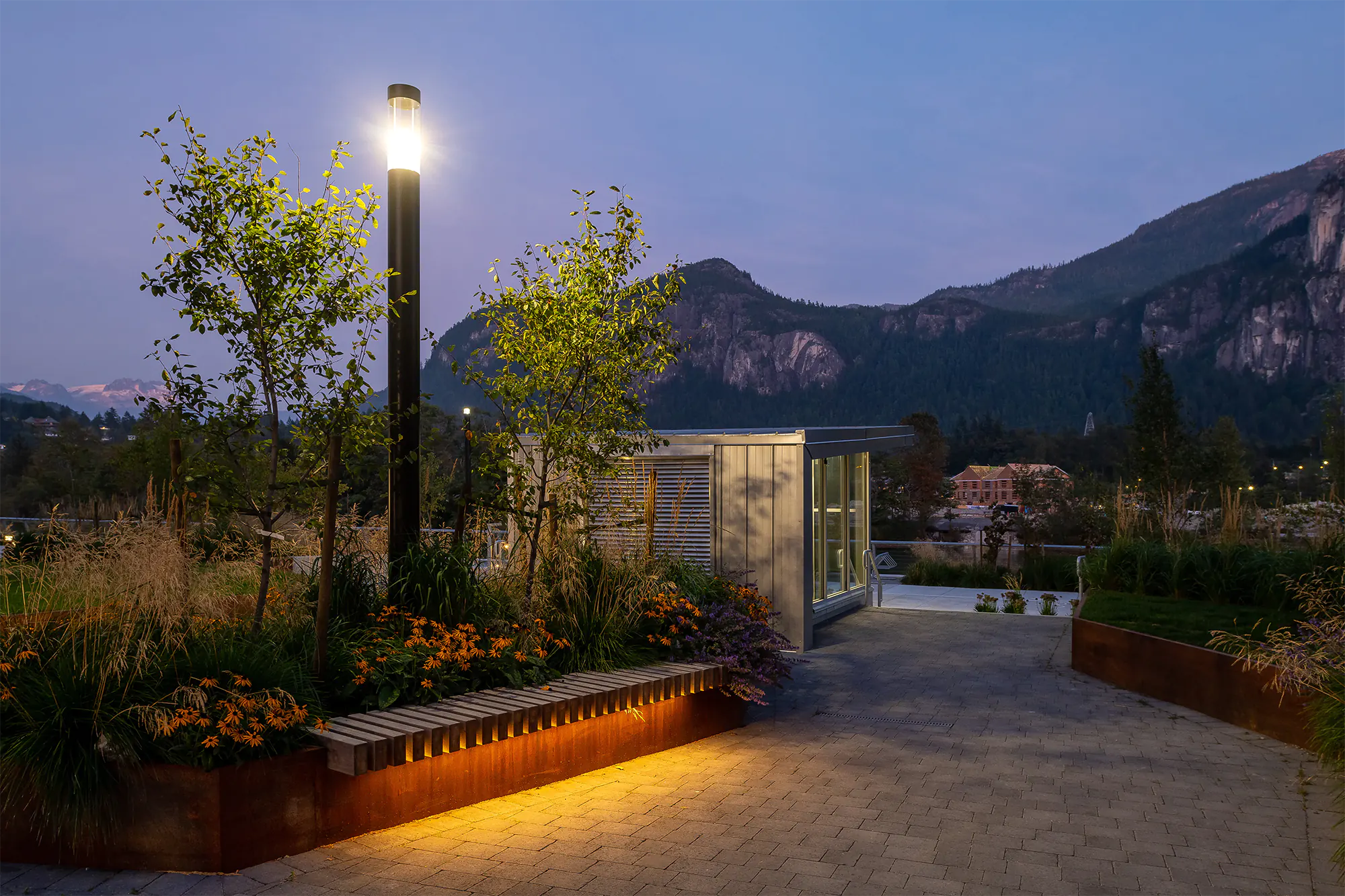 Rooftop garden at dusk featuring tall black bollard light, linear LED in corten planters with native plants, small metal pavilion, and mountain backdrop