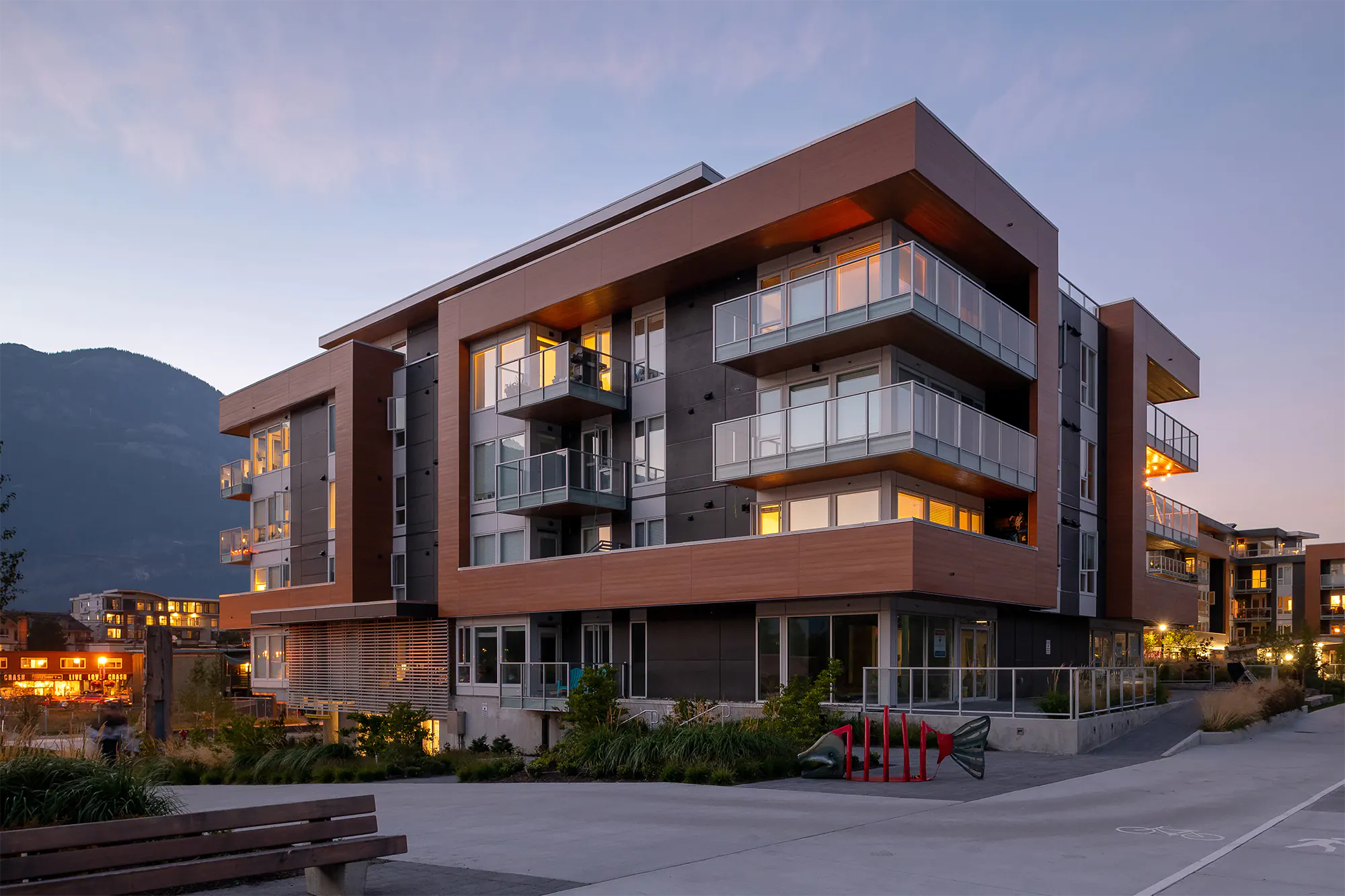 Modern multi-family building exterior at dusk with terracotta and gray wood-metal cladding, illuminated balconies, linear LED accents along edges, red fish sculpture, and mountain backdrop